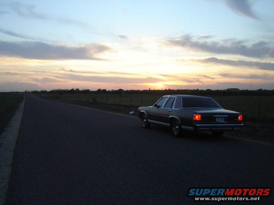 Rear view of a dark 1980s Ford sedan parked on the side of a rural road at sunset