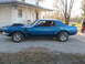 1969 Ford Mustang - A blue muscle car with a hood scoop and spoiler is parked on a gravel driveway.