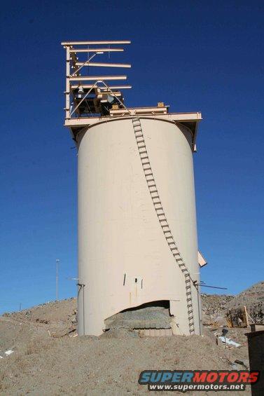 Large industrial silo with a ladder on the side, against a clear blue sky. Some damage is visible at the base