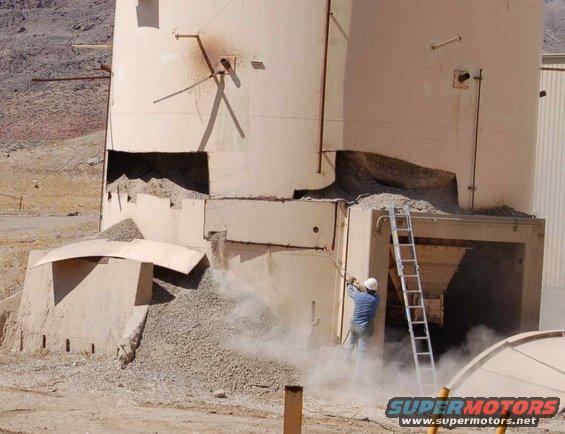 Worker on ladder at damaged industrial silo, dust rising. Supermotors.net logo visible