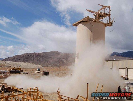 Dust cloud erupting from a tall industrial silo in a desert landscape with mountains in the background