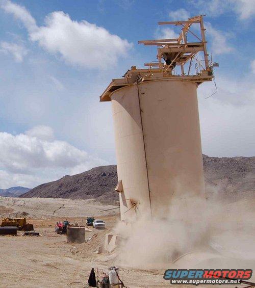 Dust cloud erupting from base of industrial silo with vehicles and machinery in background