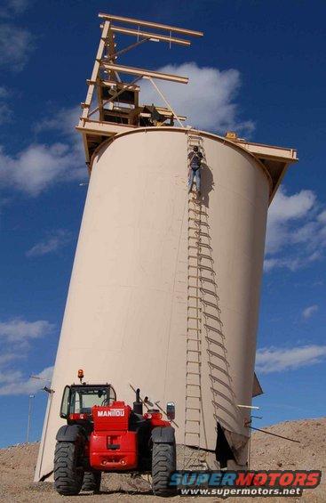 Man climbing ladder on large silo; red Manitou telehandler parked below