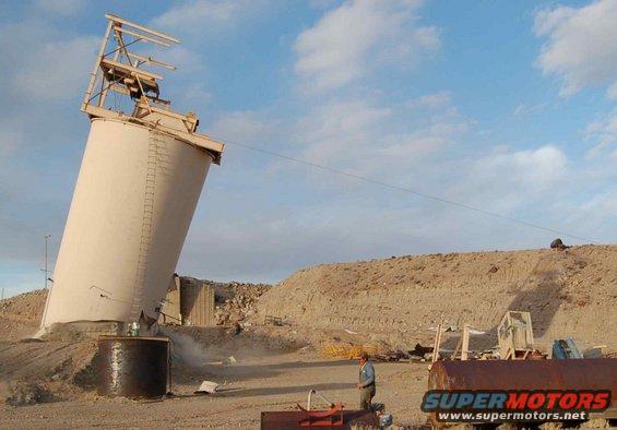 Tilted industrial silo with scaffolding against a blue sky, dusty ground, and a man standing nearby