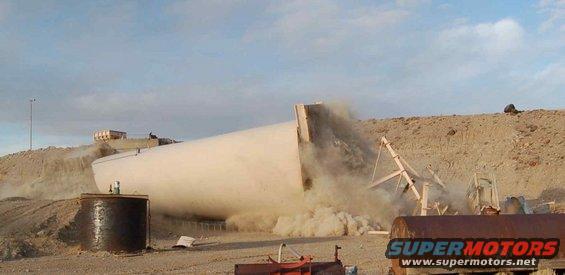 Large white cylindrical object falling in a dusty desert scene with construction equipment
