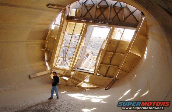 Man in jeans and dark shirt inside a large, unfinished concrete tunnel with wooden framing and windows, casting shadows
