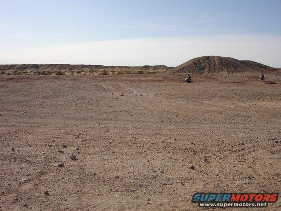 Two dirt bikes riding in a wide, flat desert area with hills in the background