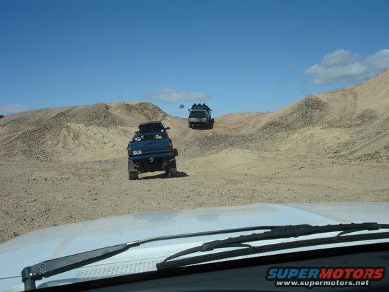 Two blue trucks off-roading in a desert with sandy hills under a clear blue sky