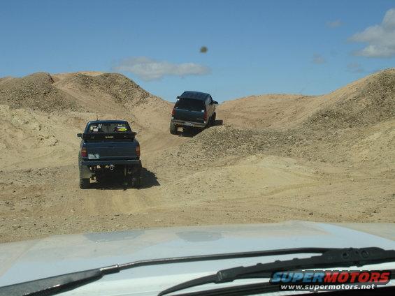 Two trucks driving on dirt hills under a blue sky. One truck is in the foreground, the other is further up a hill