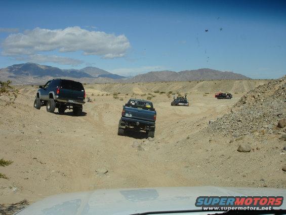 Several off-road vehicles navigate a sandy, hilly terrain under a bright blue sky with distant mountains