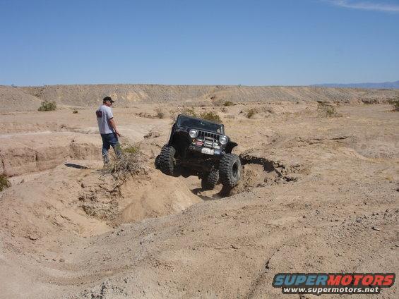 Black Jeep Wrangler navigating a steep dirt embankment in a desert landscape. A person stands nearby watching