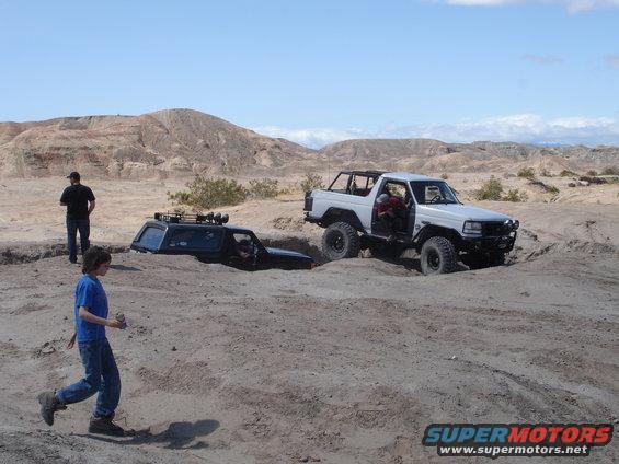 Two off-road vehicles are stuck in a sandy ditch in a desert landscape. One is white with its door open, the other is dark blue and mostly submerged