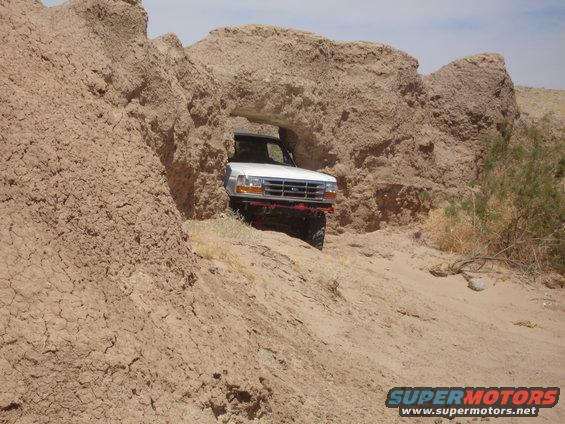 White 1996 Ford Bronco driving through a rock arch in a desert landscape