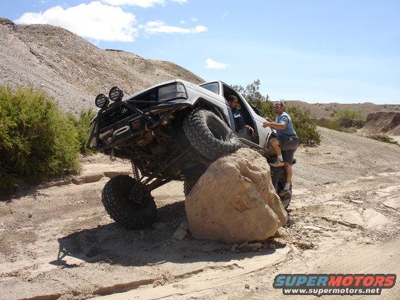White off-road SUV with a roll cage, large tires, and aftermarket bumper, climbing over a large rock