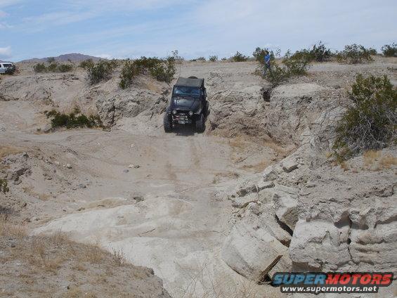 Jeep Wrangler driving down a rocky, off-road trail in a desert landscape. Another white SUV is visible in the distance