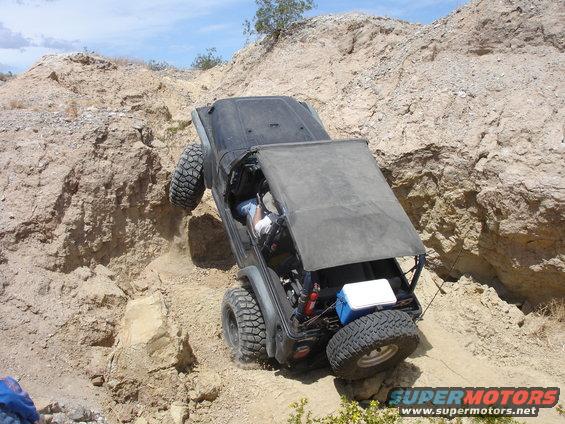 Jeep Wrangler with soft top up, tires digging into dirt on a steep incline, driver visible