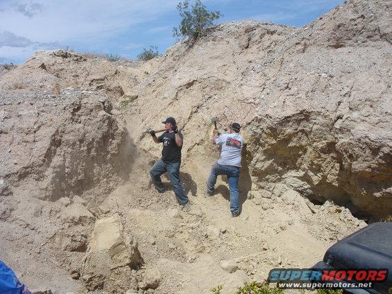 Two men with pickaxes digging into a dirt hillside under a blue sky. A black vehicle fender is visible in the lower right