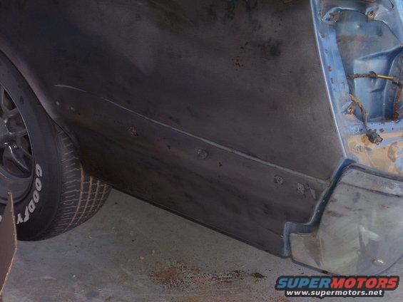 Close-up of a car's rear quarter panel being prepped for repair, showing primer and some rust spots. A tire is visible on the left