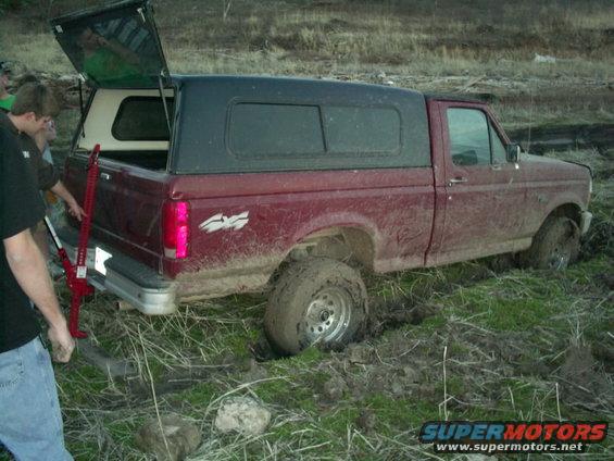 Mud-covered red pickup truck with a black camper shell stuck in a muddy field
