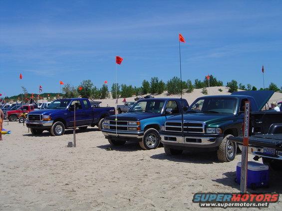 Several pickup trucks parked on a sandy beach with red flags in the background