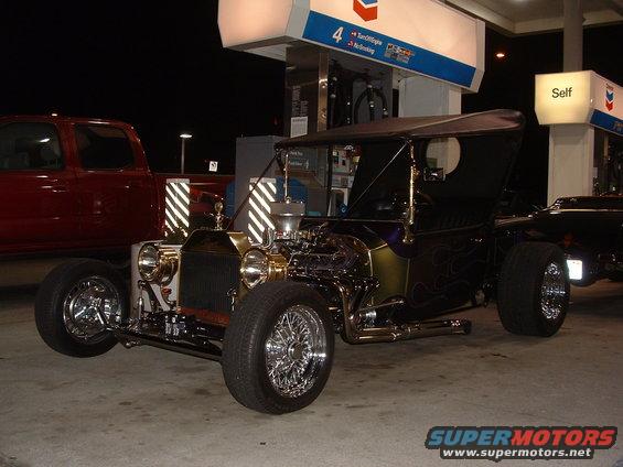Custom hot rod with exposed engine and flame paint at a gas station, with a red pickup truck in the background