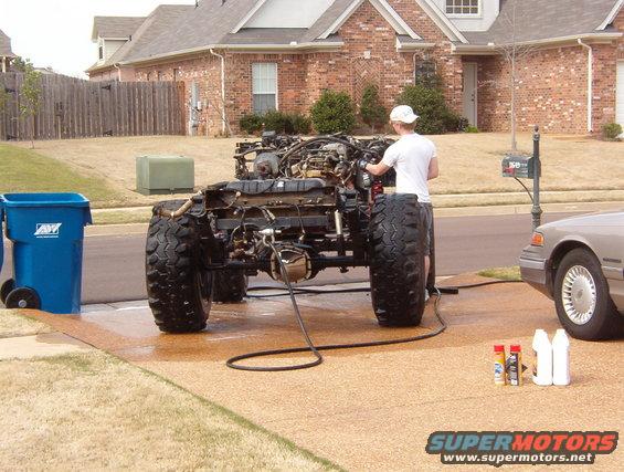 Vehicle chassis with engine and suspension visible, person working on it. Another car is parked nearby