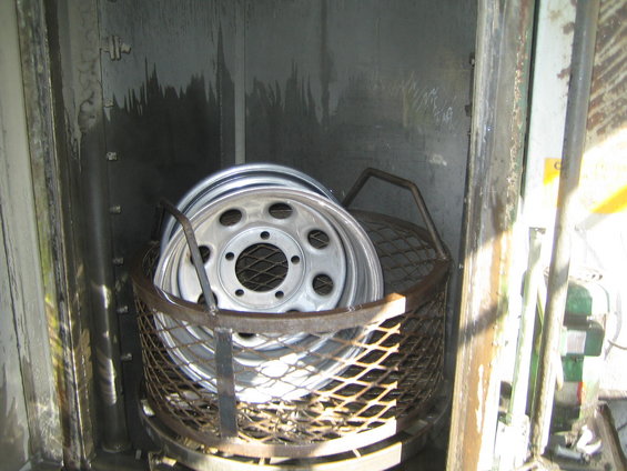 Stack of aluminum wheels with five lug holes in a metal basket inside a spray booth