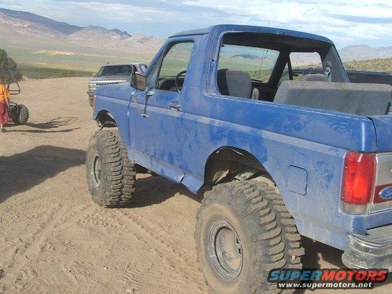 Blue Ford Bronco with large tires, seen from the rear quarter, in a sandy, outdoor environment