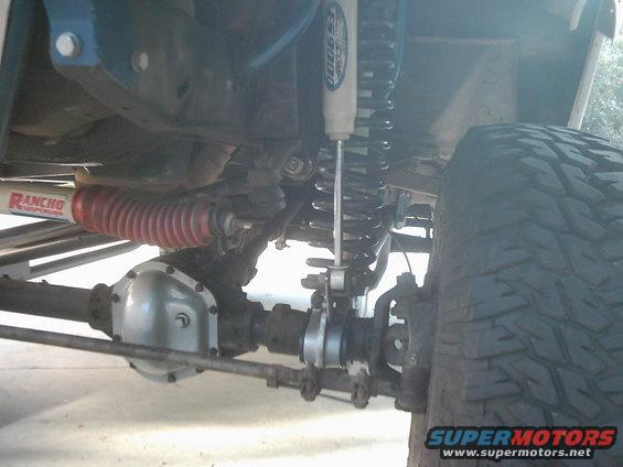 Underside of a vehicle showing a silver differential cover, Rancho steering stabilizer, and a coilover shock absorber
