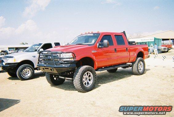 Red crew cab pickup truck with aftermarket bumper and lift kit. Another pickup is parked to its left