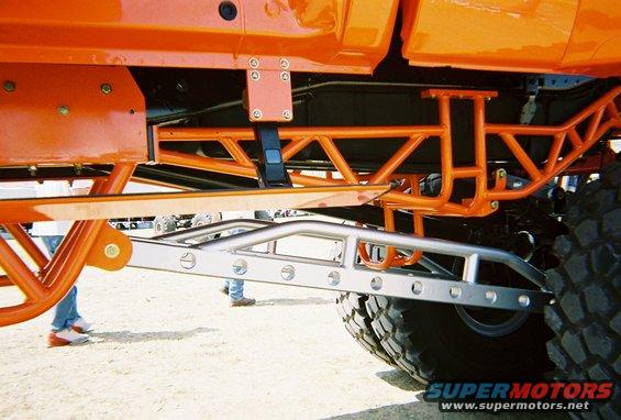 Orange custom truck frame with silver suspension components and large tires. People visible in background