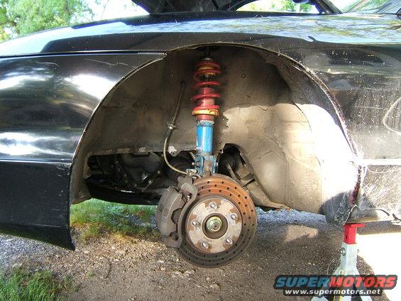 Black car's front wheel well with a blue and red coilover suspension and rusty brake rotor. The car is on a jack stand
