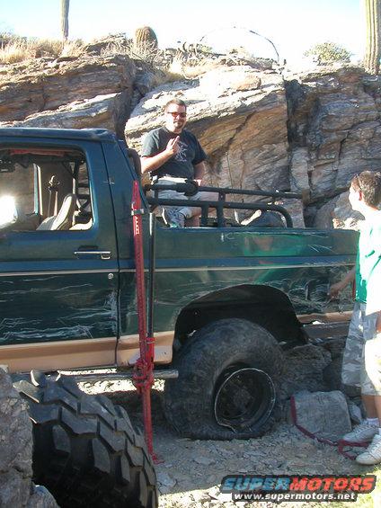 Green off-road truck with a jack on the side, two people standing around it in a rocky, desert-like area