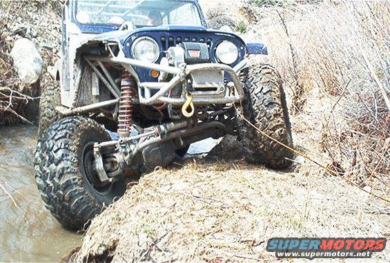 Off-road Jeep with large tires navigating a muddy stream and dirt embankment. It has a winch on the front bumper and exposed suspension