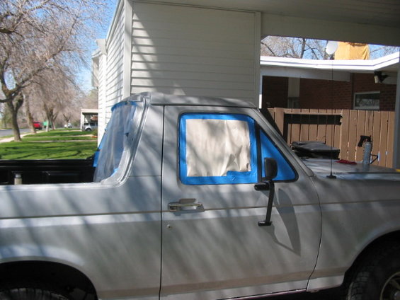Side view of a white Ford Bronco with windows taped off for painting. It's parked under a carport