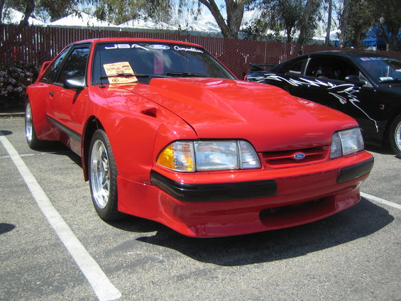 Red Ford Mustang with a big hood scoop parked in a lot. Another black car is behind it