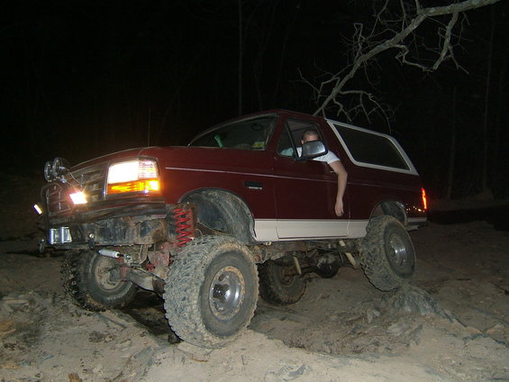 Maroon 1993 Ford Bronco with white trim, lifted suspension, and off-road tires, driving over rocks at night