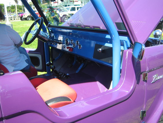 Purple Ford Bronco interior with blue roll cage, red and black seats, and dashboard gauges. Other vehicles visible in background