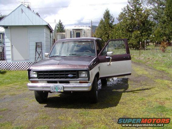 Brown and white Ford Ranger pickup truck with driver's door open, parked on a dirt road