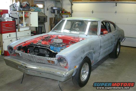 Gray primer 1970 Ford Maverick coupe with hood open, red interior, and chrome wheels in a garage