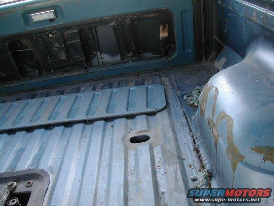 Back of a truck bed, showing the ribbed metal floor and wheel well area. Some rust and insulation are visible