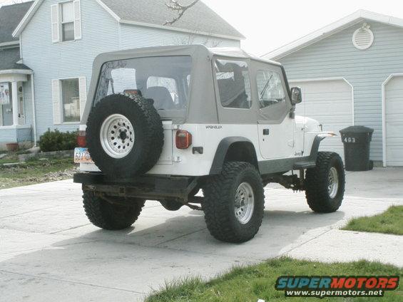 White Jeep Wrangler parked in driveway, spare tire on back. House and garage in background