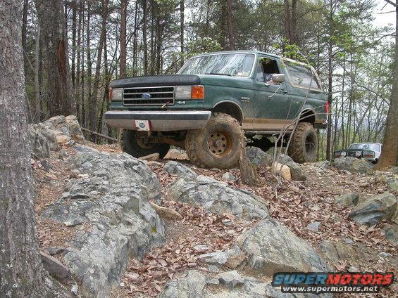 Green 1990 Ford Bronco climbing over rocks on a wooded trail. Another SUV is visible in the background