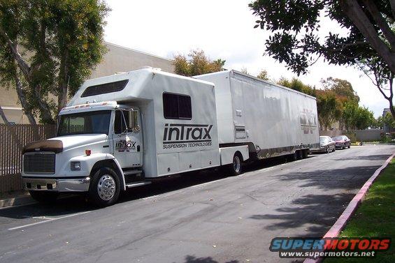 RV with a trailer, both white, with 'Intrax Suspension Technologies' logo on the side. Two cars parked in background