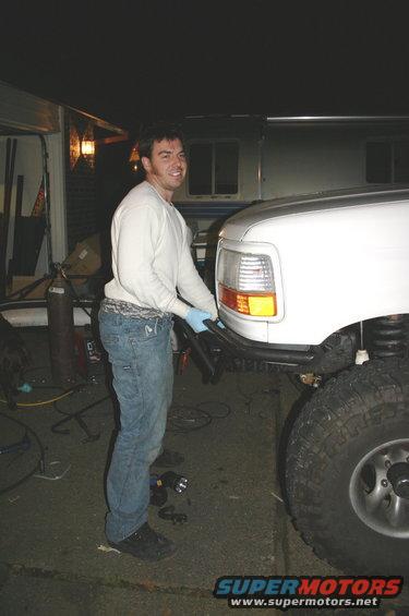 White SUV front bumper with a man in jeans and a white shirt working on it. Large tires visible