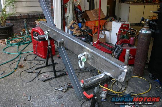 Custom steel bumper with D-ring mounts and a hitch receiver, resting on saw horses with welding equipment nearby