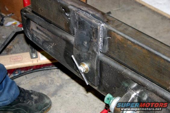 Close-up of a welded metal frame with a bolt and handle. A person's shoe is visible on the left