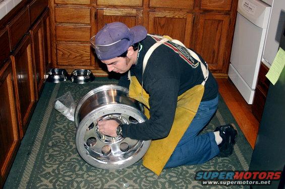Man in yellow apron and purple cap works on a large, shiny metal wheel with multiple bolt holes