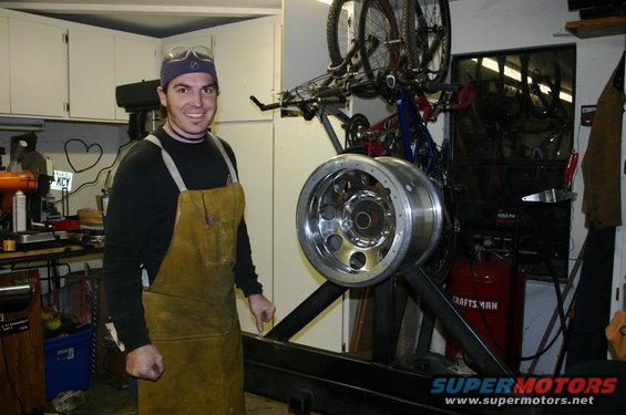 Man in workshop wearing apron and goggles, standing next to a large tire rim mounted on a stand. Bicycles are hanging on the wall behind him