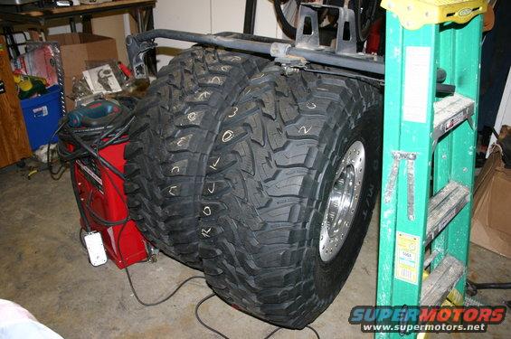 Two large off-road tires with white markings, mounted on shiny rims, next to a green ladder and red equipment